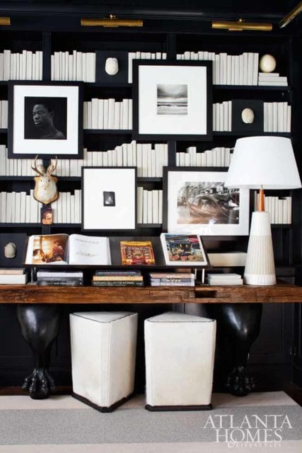 Black built-in bookshelves filled with white books, displaying framed art, a taxidermy deer head, and a table lamp. A wooden console table holds books and magazines, with two white ottomans in front.
