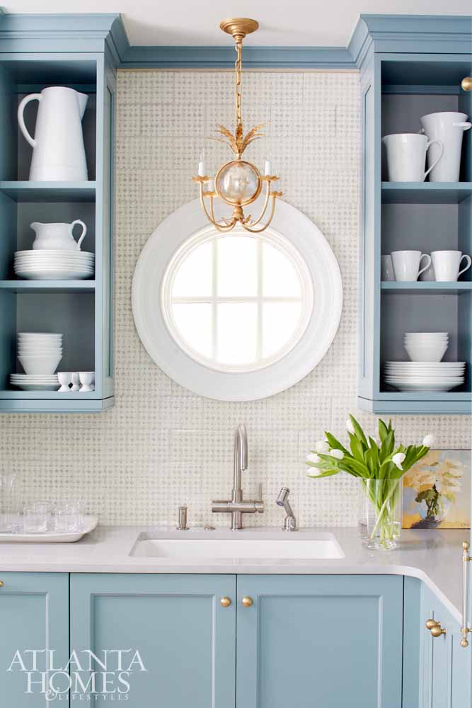 Blue cabinetry in scullery with round window over sink and white dishes in open shelving
