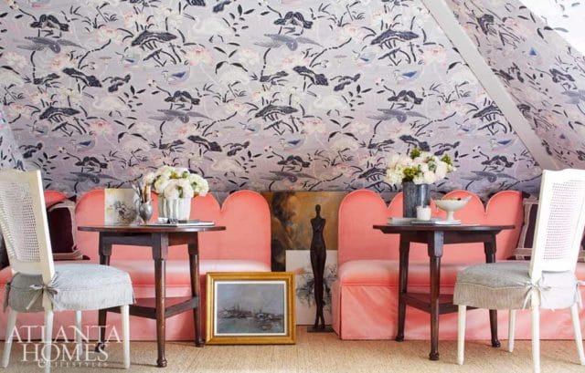 Pink scalloped sofa with cane-backed chairs and floral wallpaper. Two wooden side tables hold floral arrangements and decorative items.