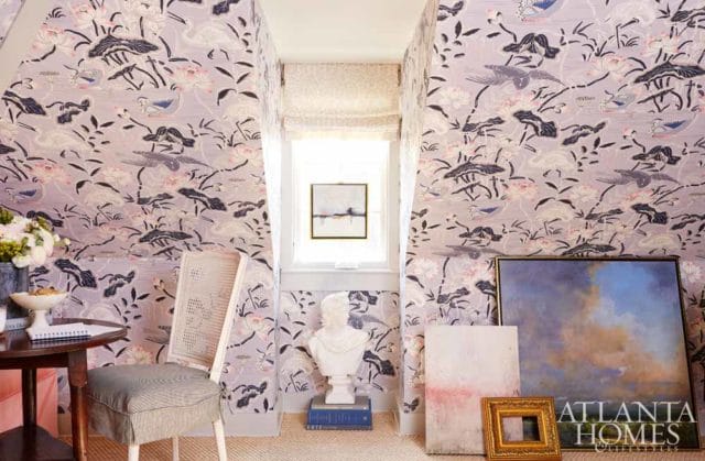 Hallway with lavender floral and bird wallpaper, a cane-backed chair with striped cushion, a round wooden table with a bowl of flowers, and artwork leaning against the wall.