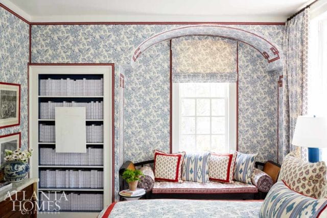 Blue and white floral wallpaper adorns walls and an arched alcove. Built-in bookshelves flank a window with a roman shade. A daybed with patterned pillows sits below the window.