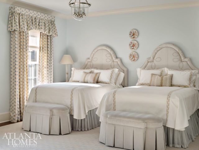 An upstairs guest room is packed with dressmaker details, including custom linens by Jacksonville, Florida’s The Linen Ladies, crewelwork lumbar pillows from Ryan Studio, a Sister Parish pattern on the window treatments and chaise longue, upholstered headboards featuring a knotted motif, and upholstered benches with skirt pleats.