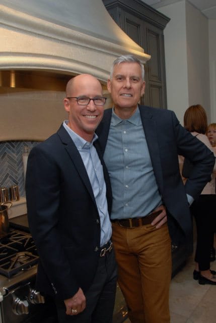 Two men smiling, one with glasses and a dark suit, the other in a blue shirt and dark suit. They are standing in front of a kitchen range and cabinetry.