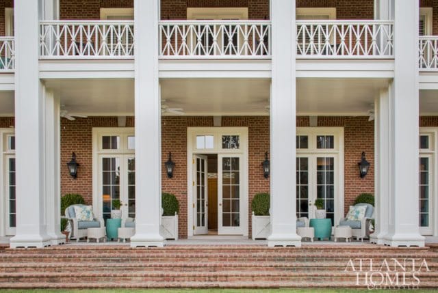 Brick house with white columns and balconies, featuring a covered porch with wicker furniture and French doors leading inside.