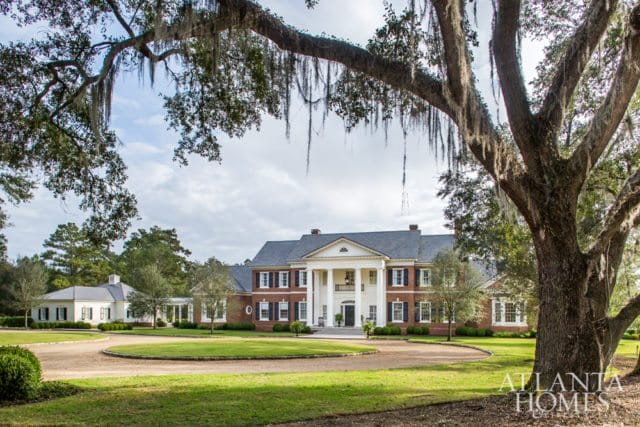 Neoclassical brick mansion with white columns, dark shutters, and a circular driveway, framed by large oak trees draped with Spanish moss.
