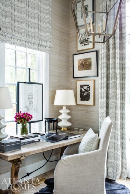 Desk area with a wooden table, white lamp, framed artwork, and a chair upholstered in light-colored fabric. The walls are covered in textured wallpaper.