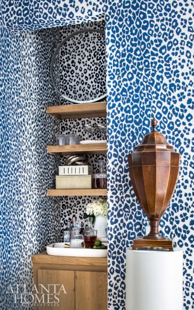 Bar area with blue and white leopard print wallpaper, wooden cabinetry, open shelves with glassware and decanters, and a decorative wooden urn on a pedestal.