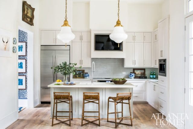 White kitchen with light wood floors, a large stainless steel refrigerator, white cabinetry, a white island with three woven stools, and brass pendant lights.