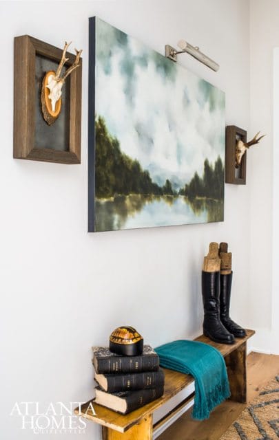Entryway with landscape painting, mounted deer skulls, stack of antique books, teal throw blanket, and black boots on wooden bench. Atlanta Homes & Lifestyles logo in corner.