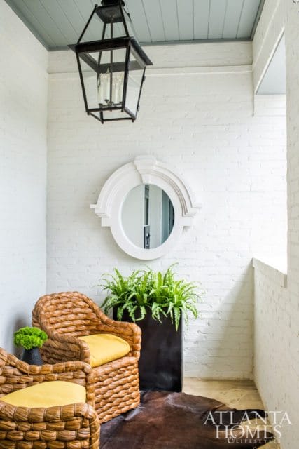 Porch with white brick walls, a black lantern, a decorative mirror, wicker chairs with yellow cushions, and a large planter filled with ferns.