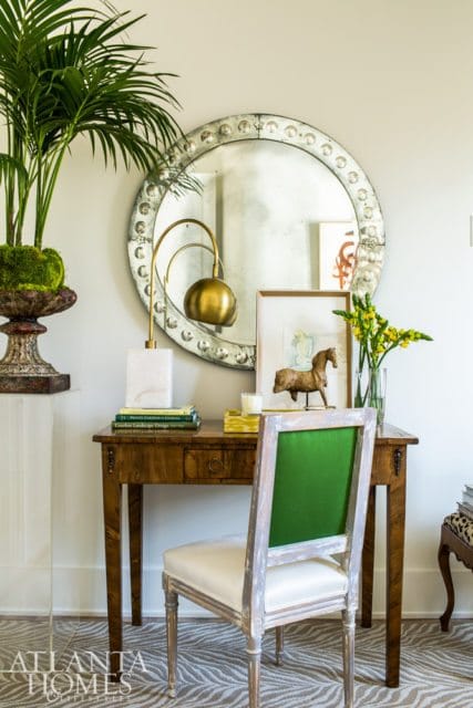 Desk with a wooden top and drawer, a chair with green upholstered back and white seat, a large round mirror with decorative frame, a lamp, books, and a plant.
