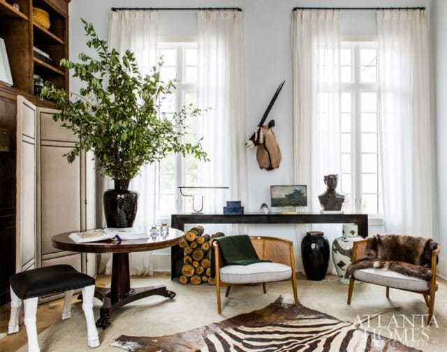 Living room with a dark wood pedestal table topped with a large black vase of greenery, two cane-backed chairs with plush cushions, and animal hide rugs on the floor.