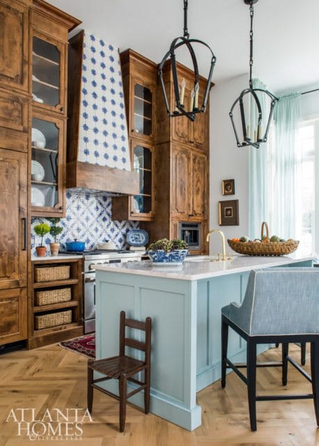 Light blue kitchen island with marble countertop and blue upholstered bar stools, surrounded by warm wood cabinetry and blue and white patterned tile backsplash.