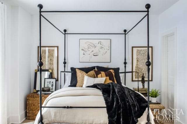 Bedroom with a black metal canopy bed, layered pillows in earth tones and black, and a black faux fur throw. Artwork hangs above the bed, flanked by framed prints.