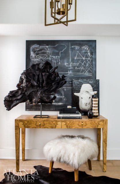 Console table with burl wood finish topped with black art, a sheep taxidermy, and decorative objects. A fluffy white stool sits in front.