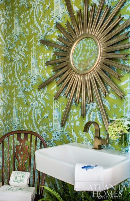 Powder room with bright green chinoiserie wallpaper, a white farmhouse sink with a brass faucet, a sunburst mirror, a wooden chair with embroidered towels, and a vase of hydrangeas.