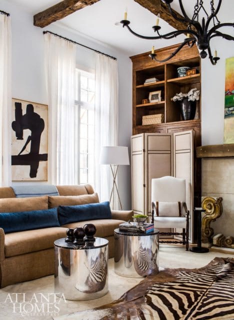 Living room with a tan sofa, blue velvet pillows, silver cylindrical side tables with zebra print, and a zebra hide rug. A wooden bookshelf and a white armchair are visible.