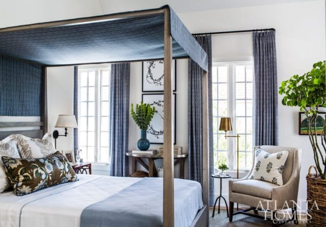 Bedroom with a four-poster bed draped in blue fabric, flanked by tall windows with blue patterned curtains. A beige armchair with a floral pillow sits near a potted plant.