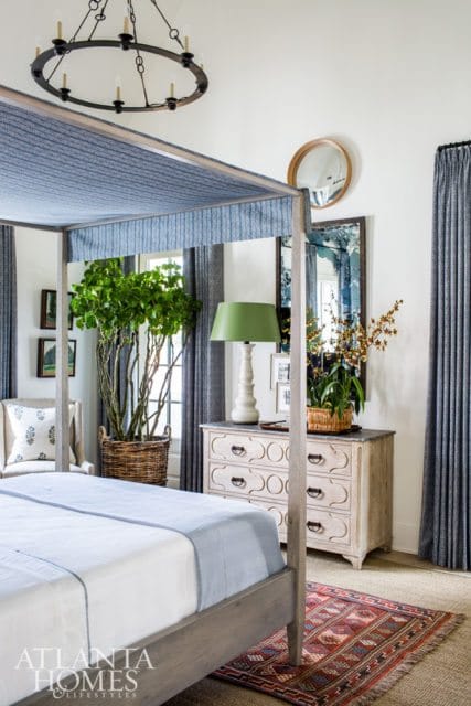 Bedroom with a gray four-poster bed, blue upholstered canopy, white bedding with blue trim, and a patterned rug. A distressed dresser with a green lamp and orchids sits nearby.