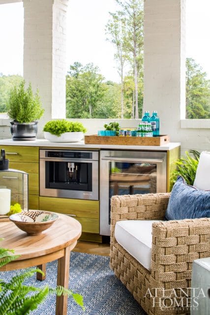 Outdoor kitchen area with lime green cabinetry, built-in coffee maker, and beverage cooler. Woven chairs and a wooden table with a blue patterned rug complete the space.