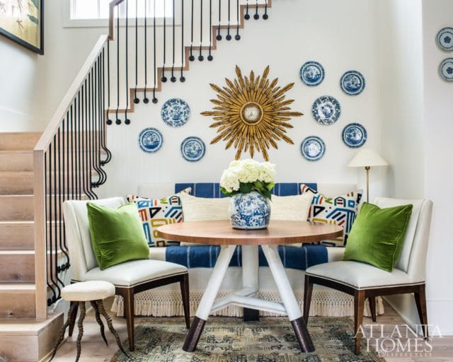 Living area with a wooden round table, two cream upholstered chairs with green velvet pillows, a bench with blue and white striped upholstery, and a collection of blue and white plates on the wall.