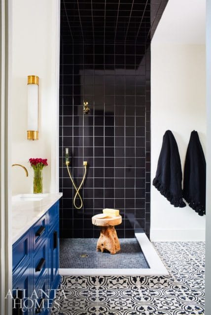 Modern bathroom with black tiled shower, blue vanity with marble countertop, and patterned floor tiles. Gold fixtures and a wooden stool add accents.