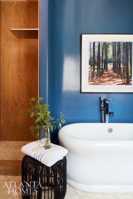 Blue bathroom with a white freestanding tub, wood-paneled closet, and framed artwork of a forest path.