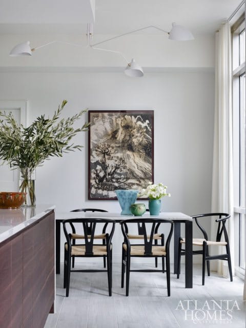 Dining area with a marble-top table, black wishbone chairs, and abstract art on the wall. A modern white chandelier hangs overhead.
