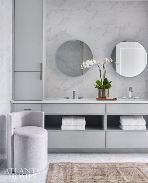 Gray bathroom with marble tile walls, double vanity with white countertops, two round backlit mirrors, and a textured gray swivel stool.