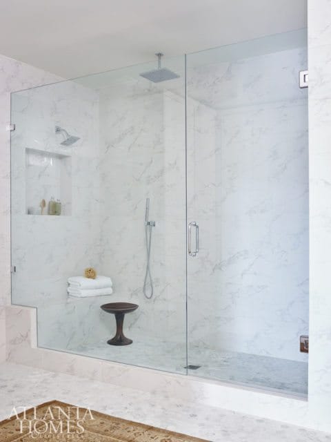 Modern bathroom with a large glass-enclosed shower featuring marble tile walls, a rain shower head, and a built-in bench. A small wooden stool sits inside the shower.