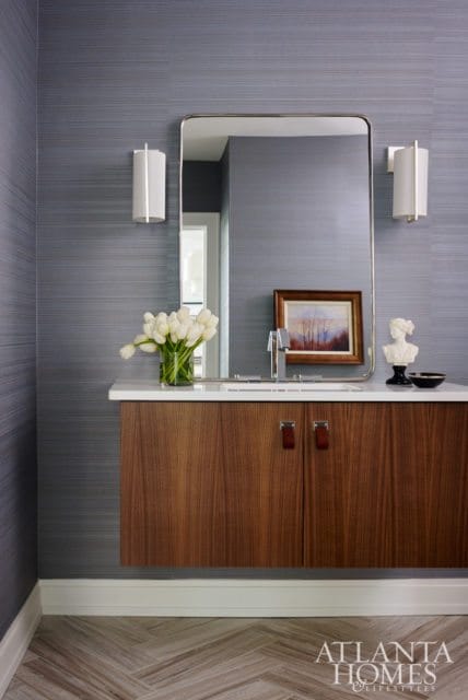 Modern bathroom vanity with dark wood cabinets, white countertop, and a large rectangular mirror. Blue textured wallpaper and sconces adorn the walls.