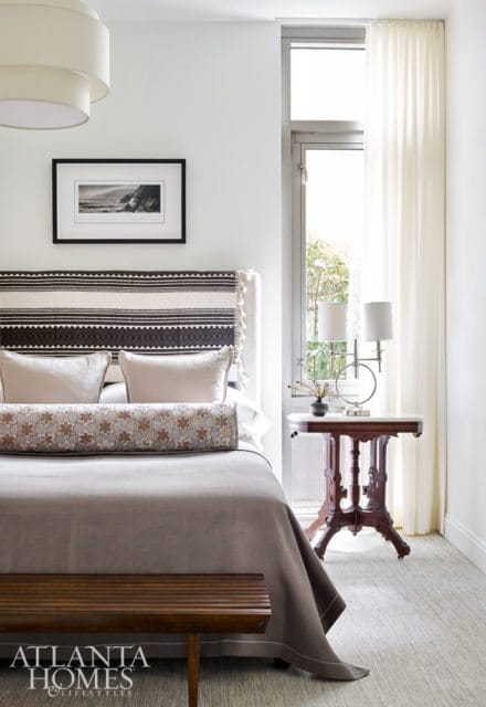 Bedroom with a king-size bed featuring a patterned headboard, neutral linens, and a bolster pillow. A framed black and white photograph hangs above the bed. A small wooden bench sits at the foot of the bed.