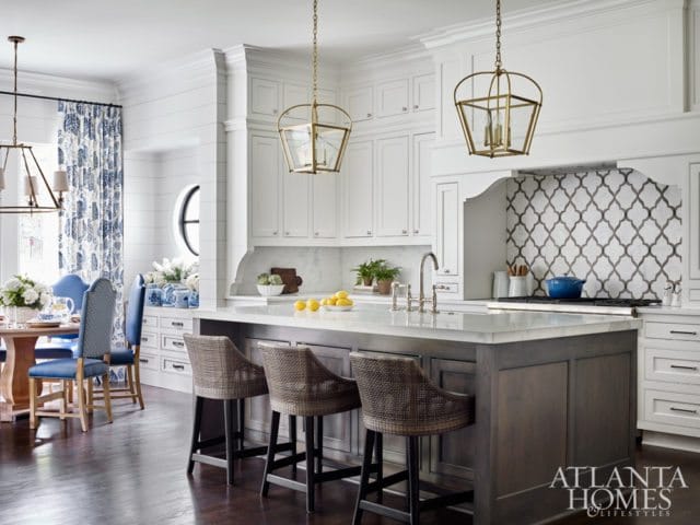 White kitchen with a dark wood island topped with marble, rattan bar stools, and a geometric tile backsplash. A dining area with blue chairs is visible to the left.