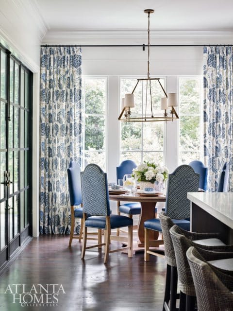 Dining room with blue patterned chairs around a round wooden table, blue floral curtains, and a brass chandelier.