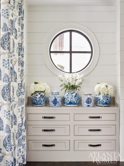 Gray dresser with black leather drawer pulls beneath a round window and shiplap walls, adorned with blue and white ginger jars and white hydrangeas.