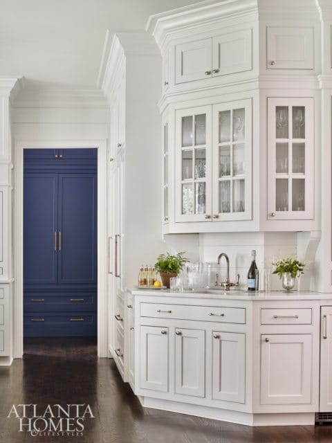 White kitchen with white shaker cabinets, glass-front upper cabinets, marble countertop, and a sink. A doorway reveals a navy blue pantry with brass hardware.