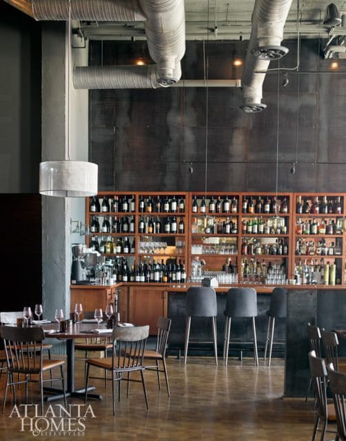 Restaurant interior with a large wine display, bar seating, and dining tables. Exposed ductwork and industrial metal accents contrast with warm wood finishes.