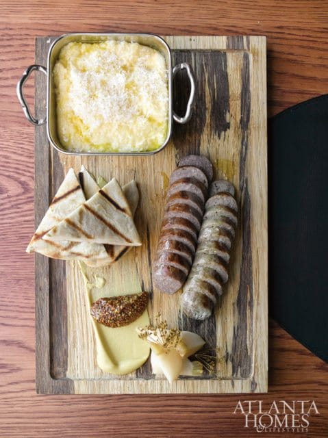 Wooden board with sliced sausages, grilled pita bread, mustard, and a bowl of polenta topped with grated cheese.