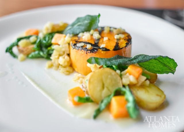 Grilled butternut squash slice topped with diced apples and herbs, served with potatoes, carrots, and greens on a white plate with a smear of sauce.