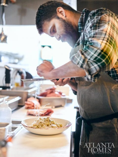 Chef grating cheese over a dish of pasta in a commercial kitchen, with raw meat in the background. Atlanta Homes & Lifestyles logo in the bottom right.