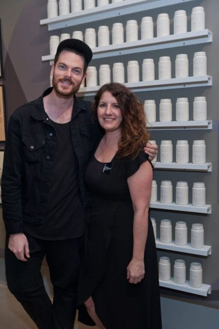 Man in black denim jacket and woman in black dress pose in front of shelves filled with white jars.