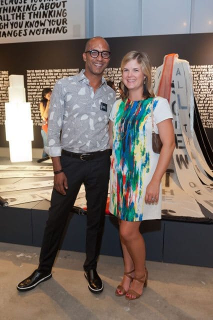 Man in a patterned button-down shirt and black pants smiles next to a woman in a white dress with a colorful abstract print.