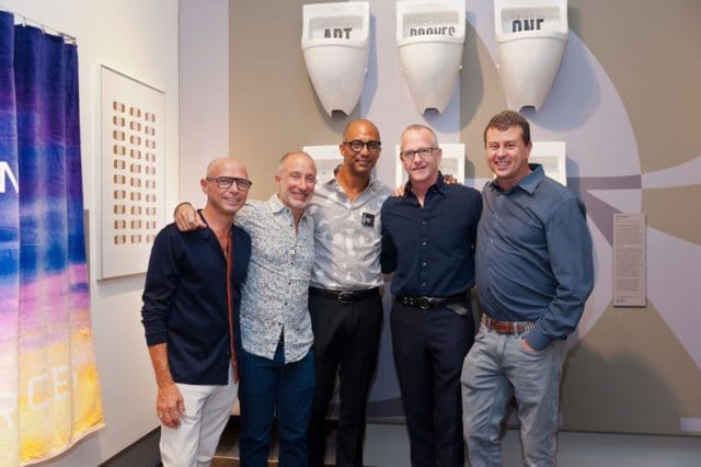 Five men stand in front of a wall with white urinals labeled "ART," "NATURE," and "OUR." To the left is a colorful tapestry and framed artwork.