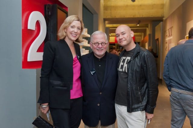 Woman in black blazer and pink top, man in navy blazer, and man in black leather jacket posing for a photo in front of a large white number 2 on a red background.