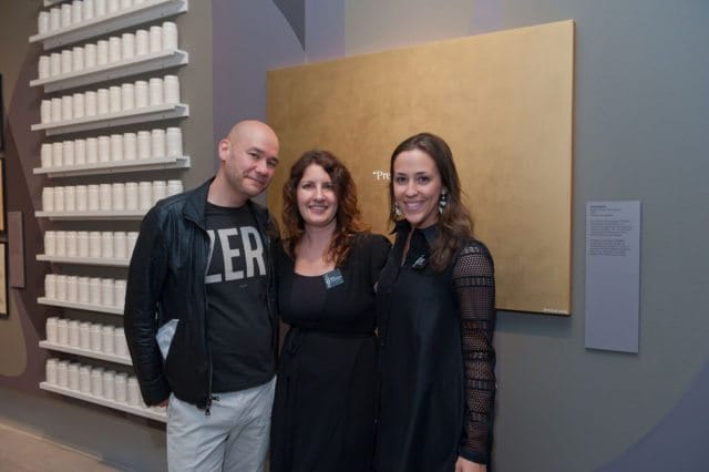 Three people stand in front of a gold artwork and a wall display of white jars. The person on the left wears a black jacket over a black t-shirt and light pants. The middle person wears a black dress. The person on the right wears a black dress with mesh sleeves.