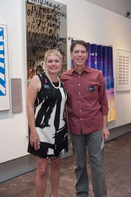 A woman in a black and white dress and a man in a red patterned shirt stand together in front of artwork. Both are smiling and wearing name tags.