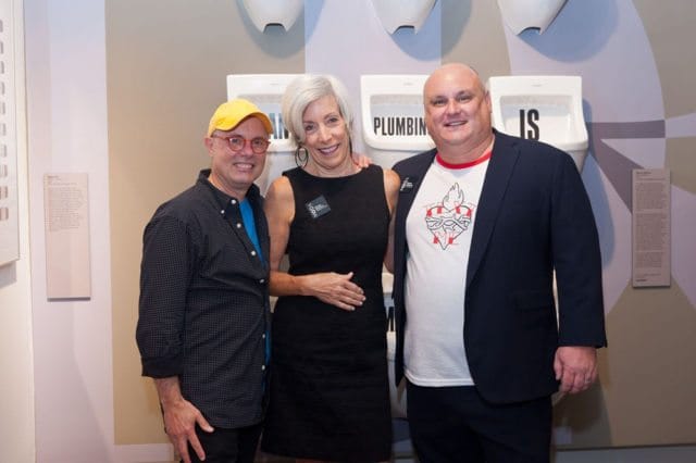 Three people pose in front of urinals with text that reads "PLUMBING IS".