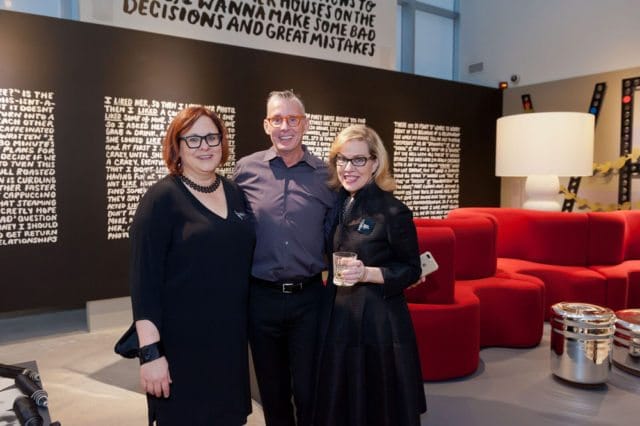 Three people pose in front of a black wall with white text. The person on the left wears a black dress and necklace. The person in the middle wears a grey shirt and glasses. The person on the right wears a black jacket and glasses, holding a drink.