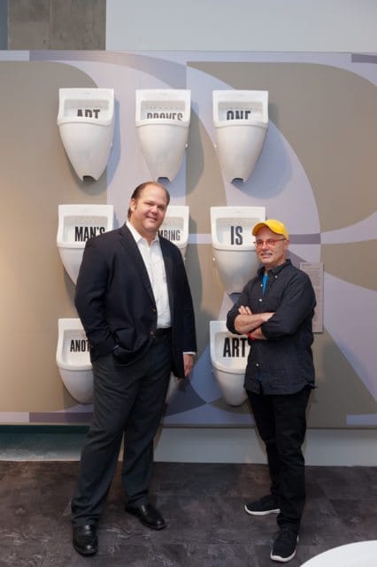 Two men stand in front of a wall display of white urinals with text. The man on the left wears a dark suit jacket over a white shirt and dark pants. The man on the right wears a dark patterned shirt, black pants, and a yellow cap.