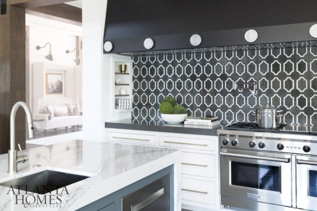 Modern kitchen with a geometric black and white tile backsplash, stainless steel range, and marble island with a waterfall edge. A glimpse of a living area is visible through an opening.